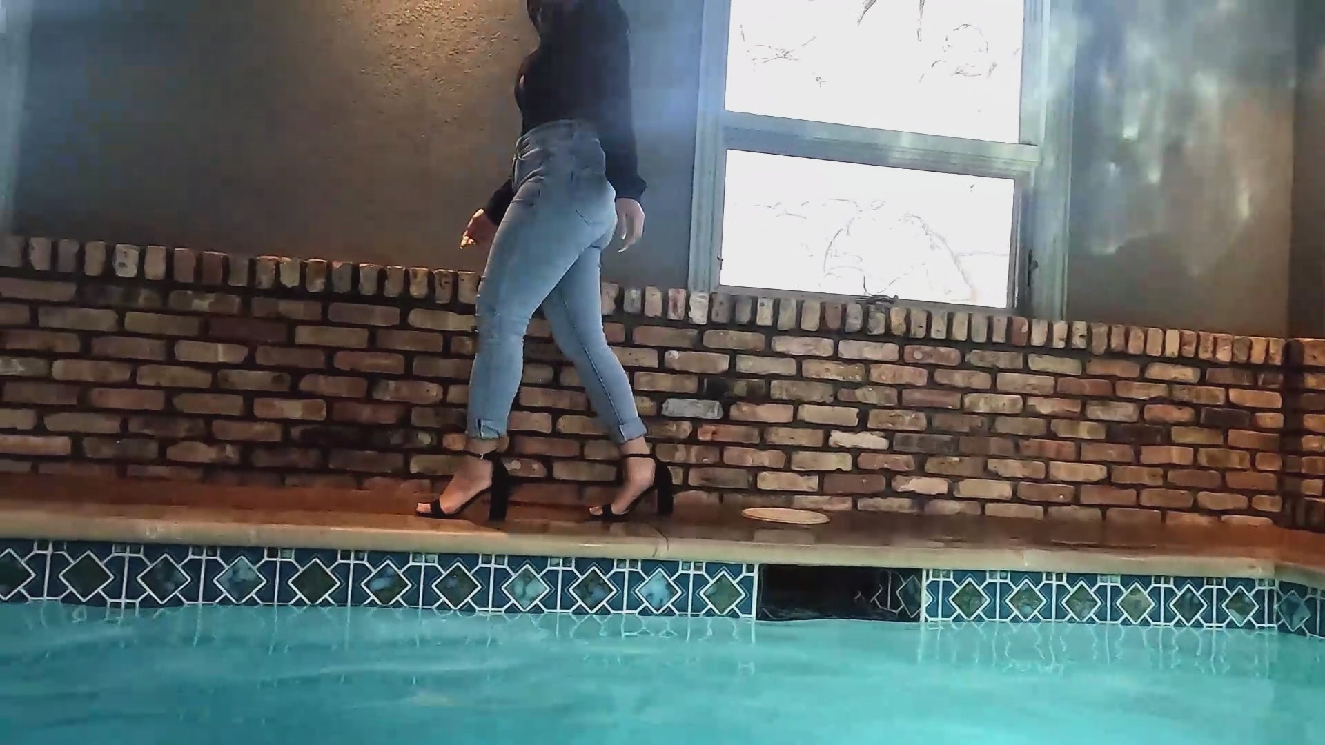 Reyna springt im Jeans und glänzenden schwarzen Oberteil in den Pool