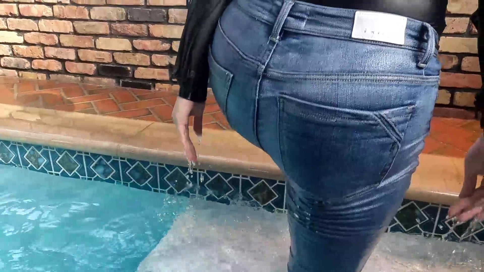Reyna springt im Jeans und glänzenden schwarzen Oberteil in den Pool